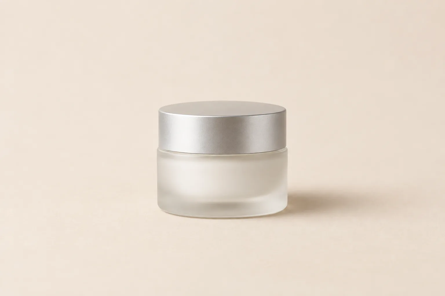 Small unbranded frosted glass eye cream jar with a matte silver lid on a sand background